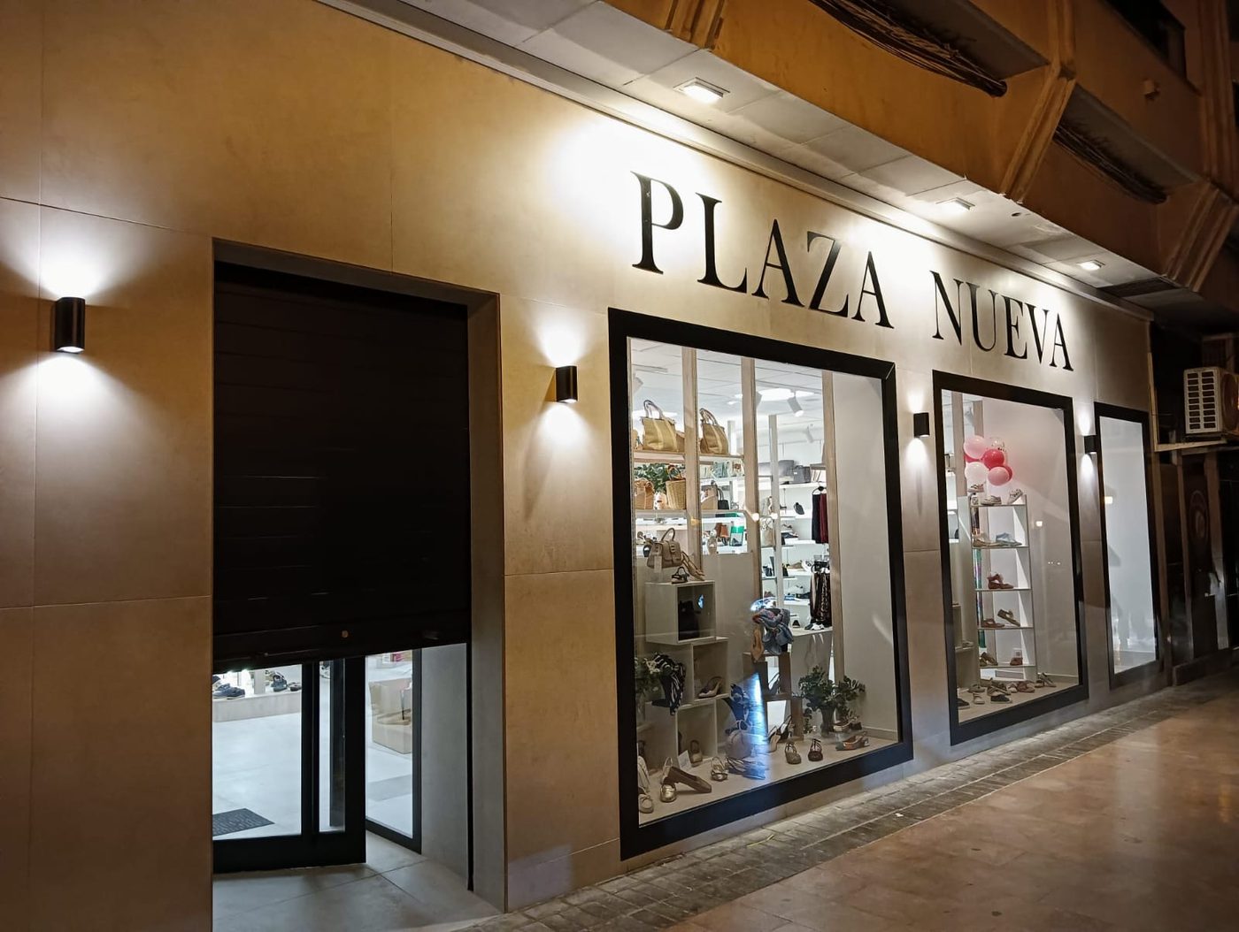 Fachada de la zapatería Plaza Nueva iluminada, con dos escaparates y letras negras sobre piedra clara